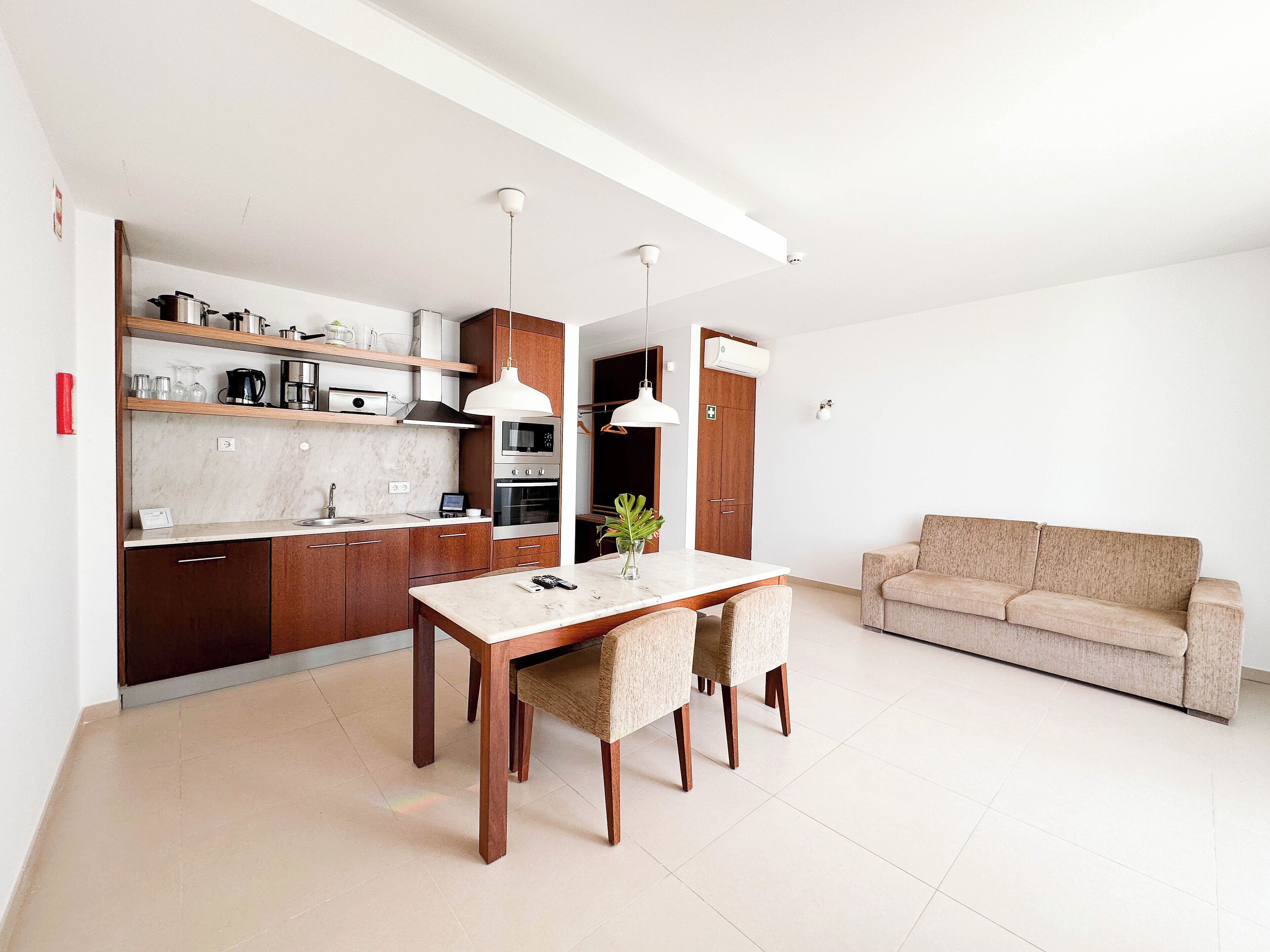 A modern kitchen with wooden cabinets, a marble countertop, and stainless steel appliances. There is a dining table with chairs and a beige sofa in the living area.