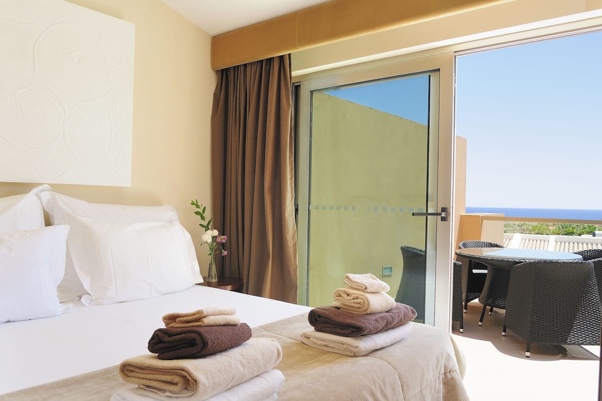 Una habitació d'hotel amb un llit, un balcó amb taula i cadires, i vista al mar.