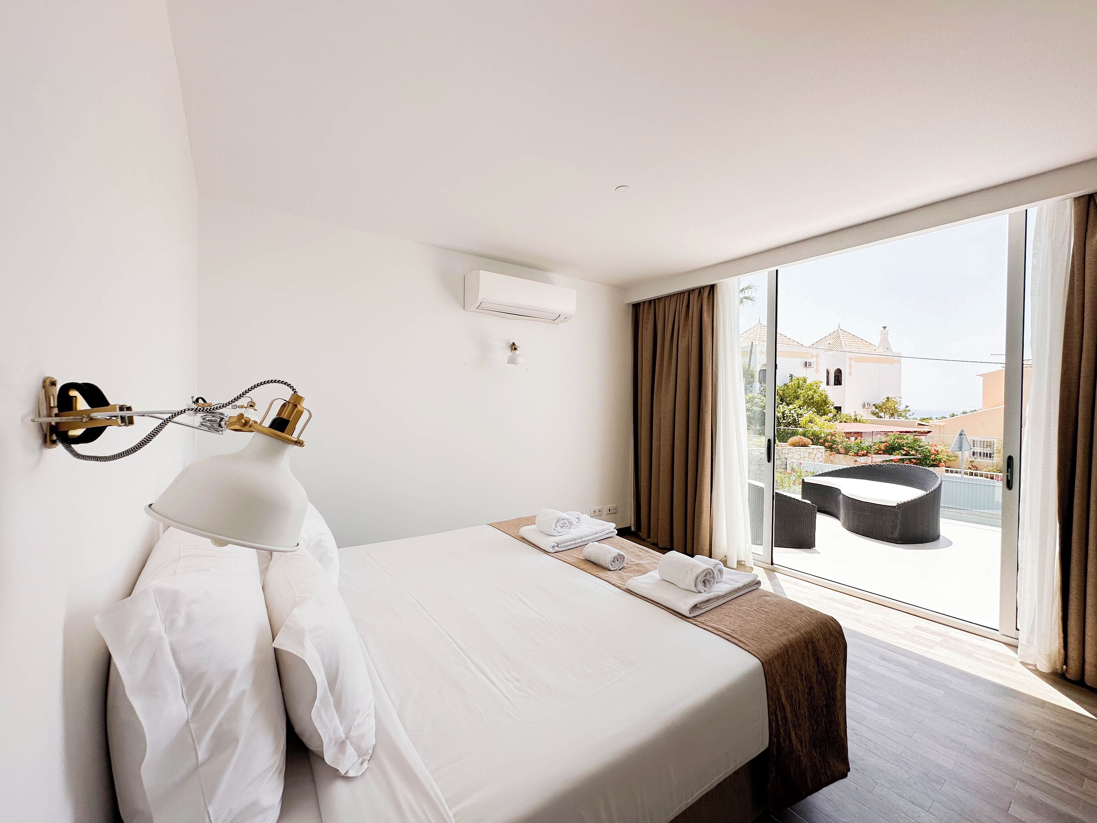 A hotel room with a large bed, a bedside table with a lamp, a view of the outside balcony, and a window with curtains.