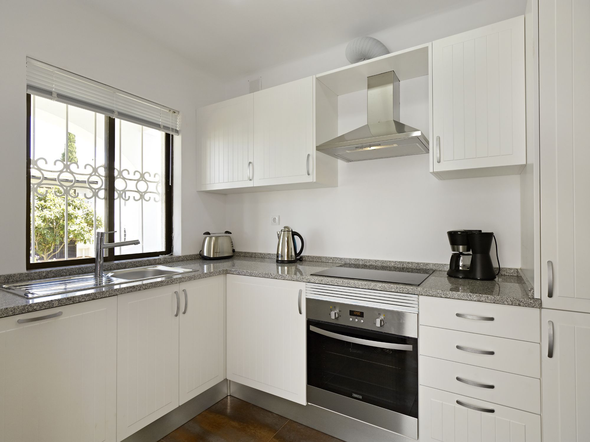 A modern kitchen with white cabinetry, a stainless steel range hood, a built-in oven, a coffee maker, and a window with decorative ironwork.