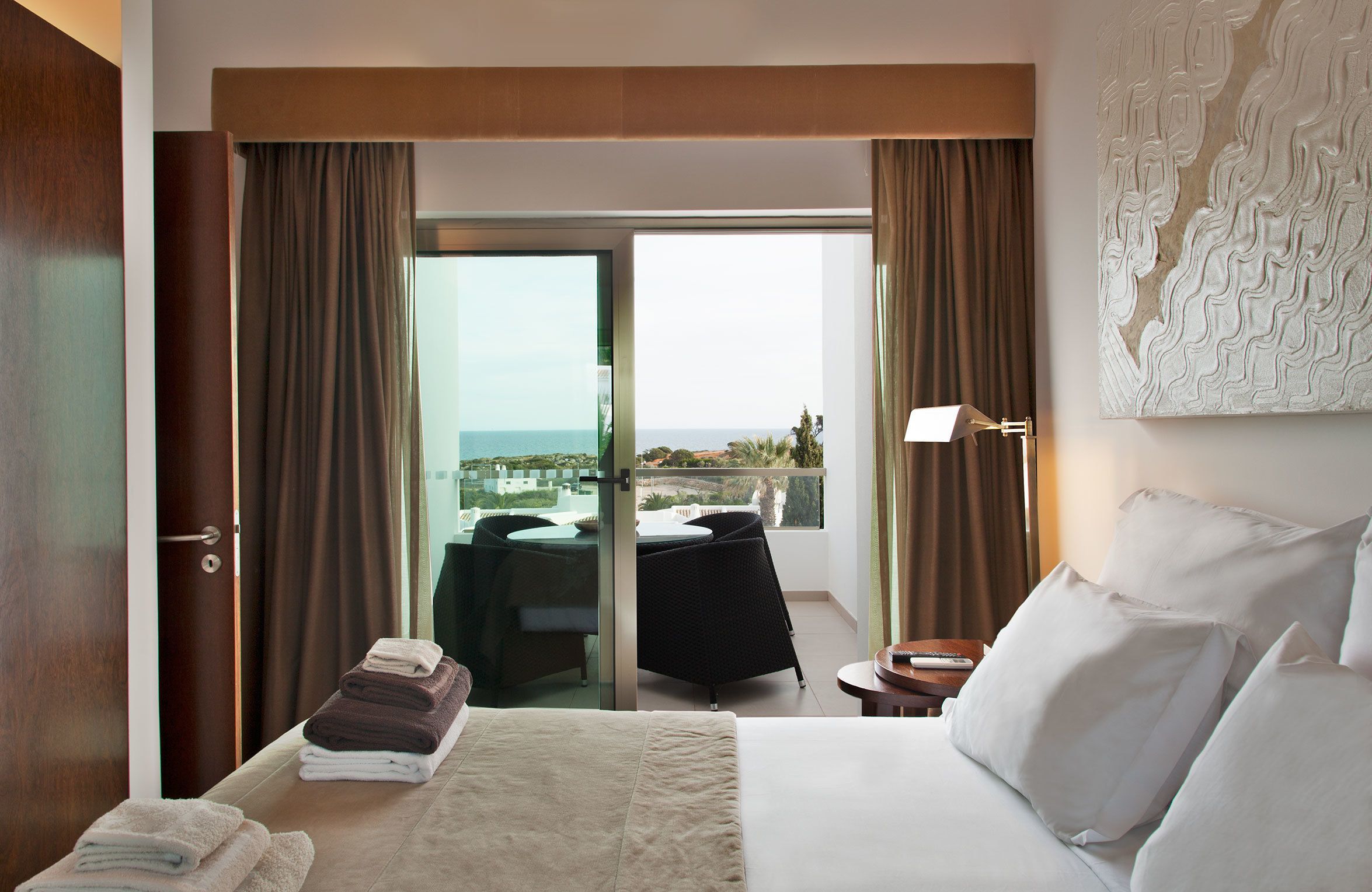 A hotel room with a bed, a table with a chair, a balcony with a view, and a door.