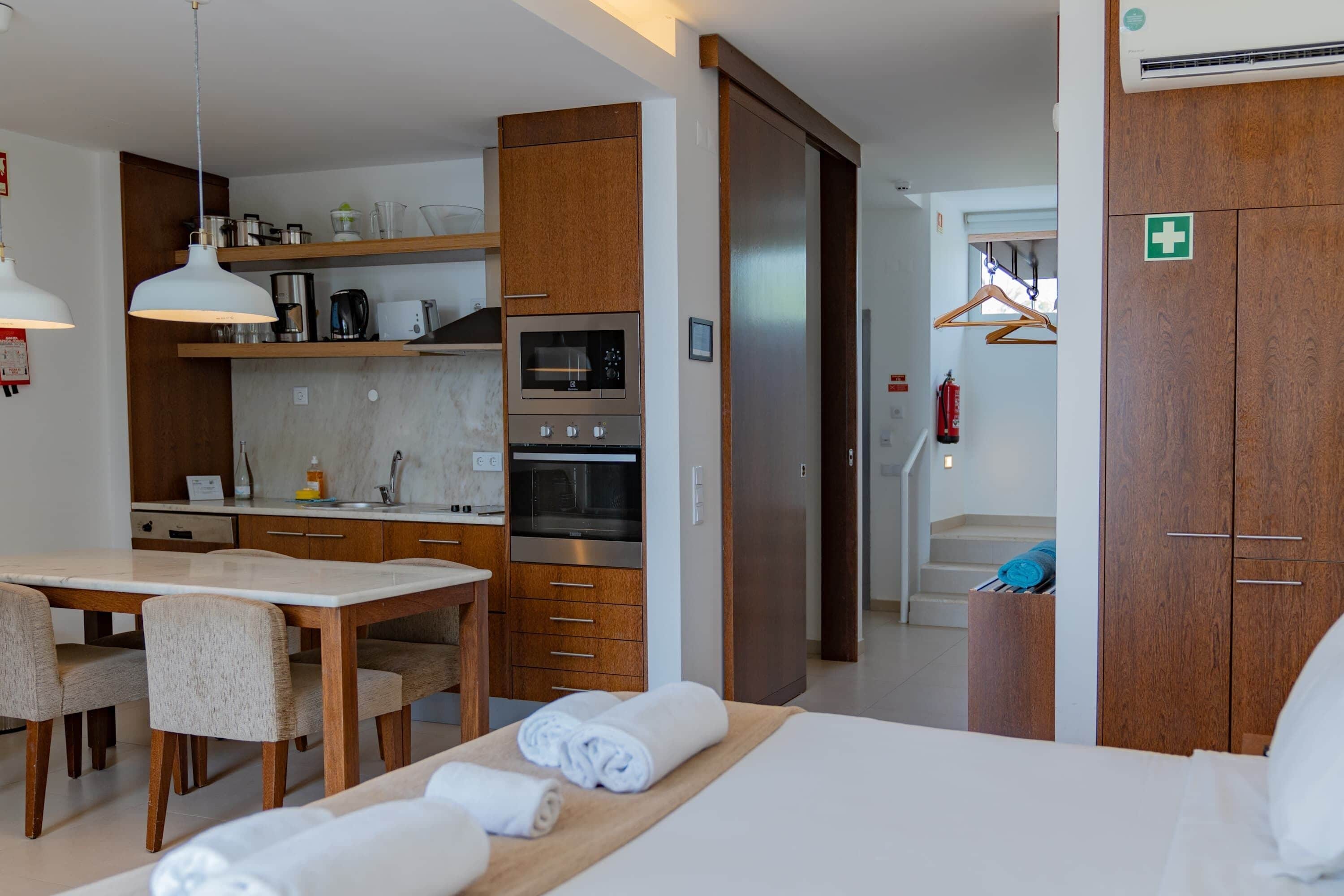 A modern hotel room with a wooden dining table, a bed with white linens, a built-in oven, and a wooden wardrobe.