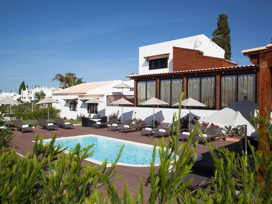Un espace piscine agrémenté de chaises longues et de parasols, entouré de bâtiments blancs dotés de grandes fenêtres et d’une terrasse en bois.