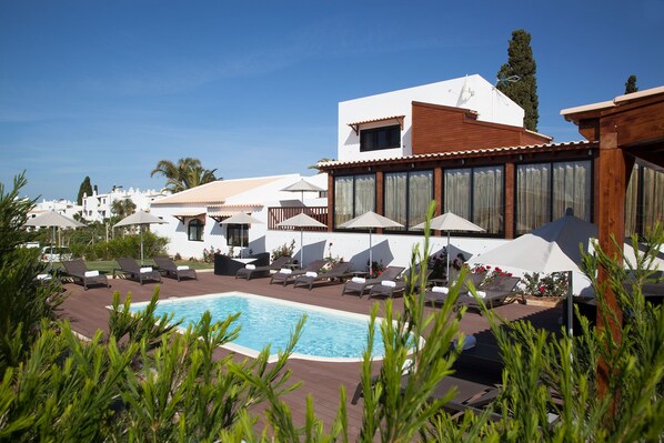 A pool area with lounge chairs and umbrellas, surrounded by white buildings with large windows and a wooden deck.
