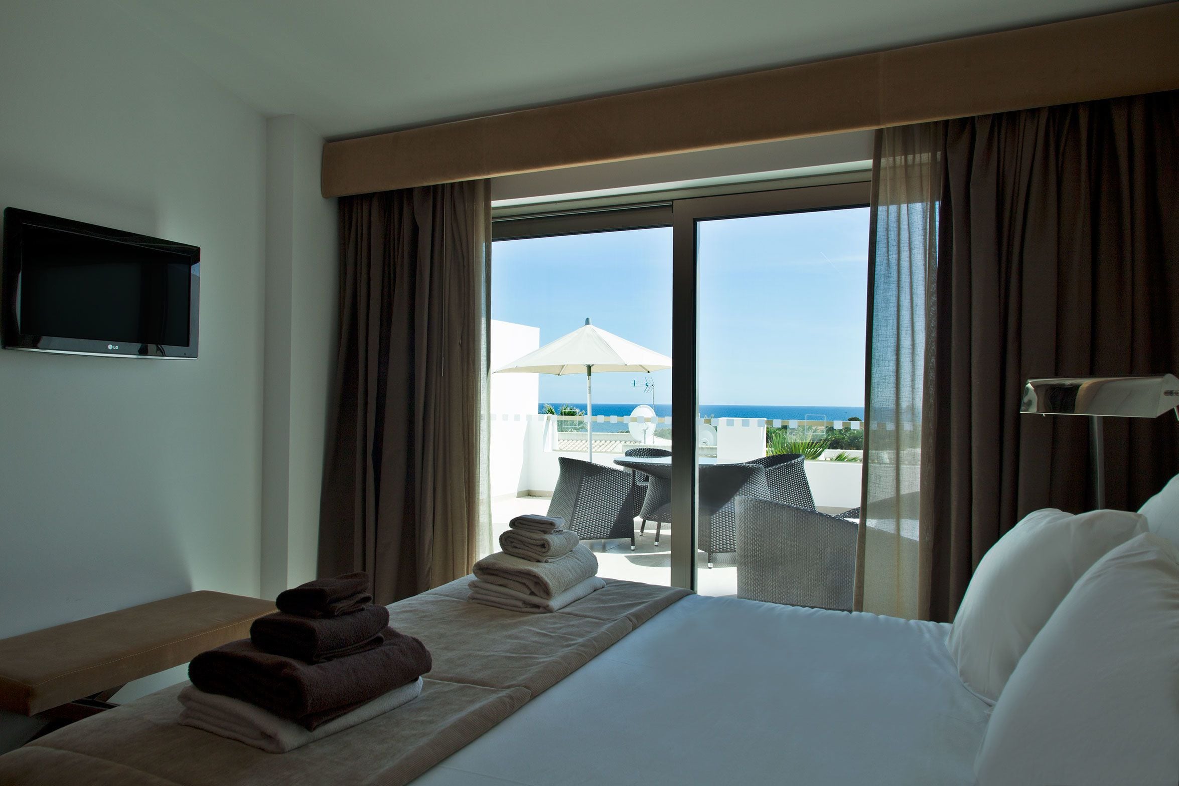 A hotel room with a bed, a TV, a view of the sea, and a balcony with outdoor furniture.