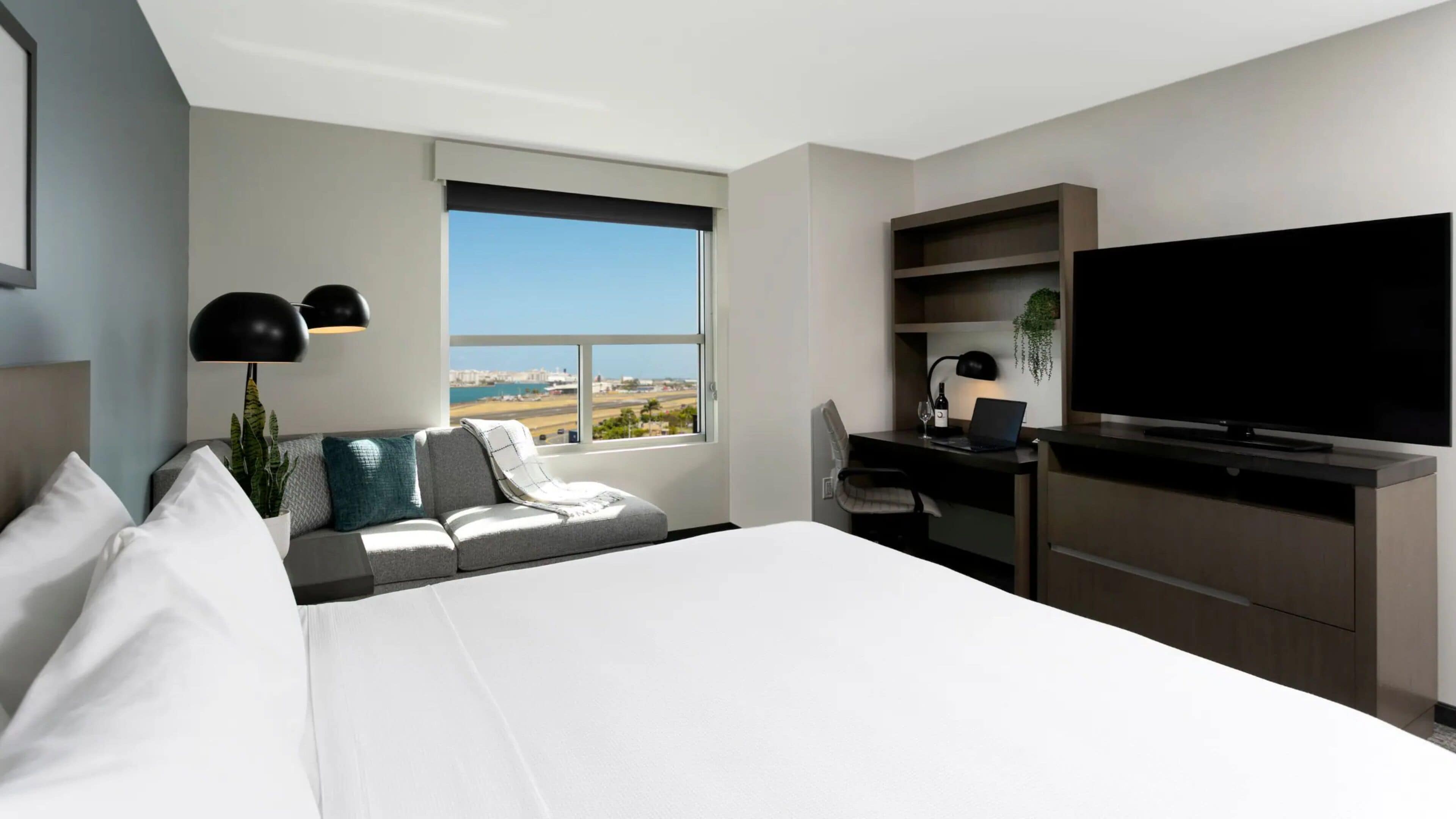 A modern hotel room with a large bed, a desk with a laptop, a TV, and a view of the cityscape.