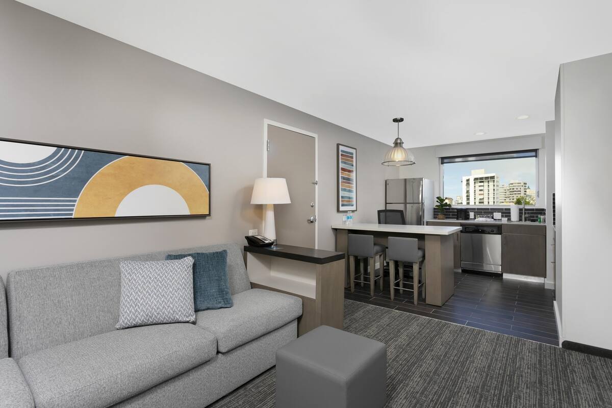 Een moderne hotelkamer met een grijze bank, een klein tafeltje, een lamp, een kitchenette en een groot raam met uitzicht op de stad.