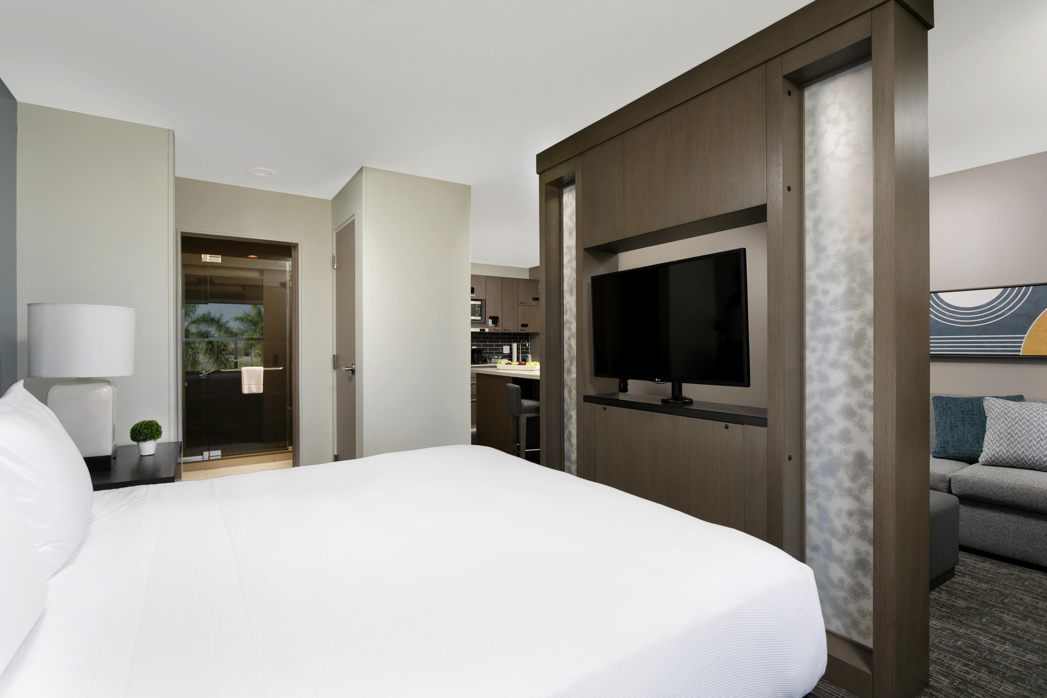 A hotel room with a large bed, a TV, a sofa, a desk, and a bathroom visible through an open door.