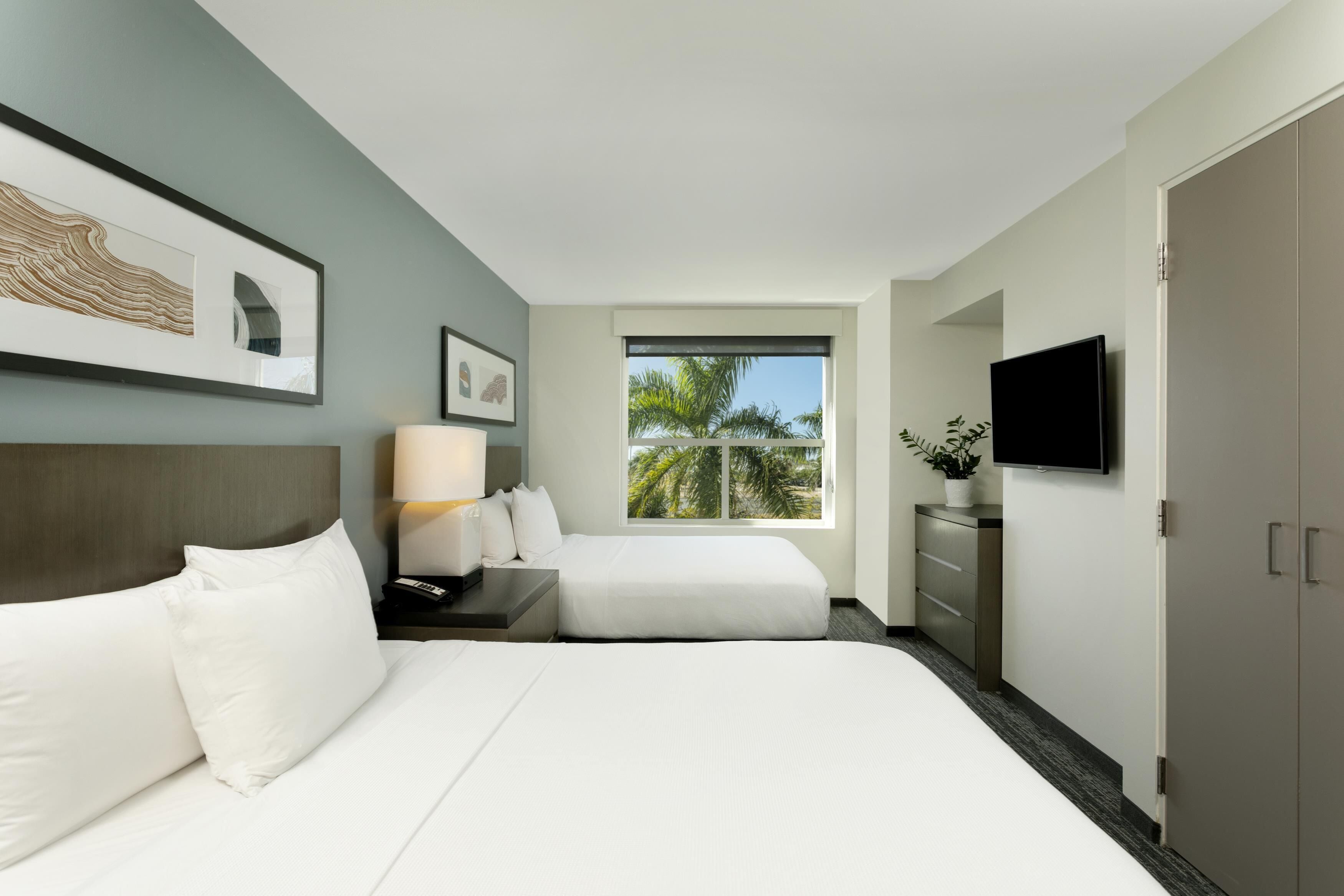 Habitación de hotel con cama, escritorio, lámpara, televisor, armario y ventana con vista a palmeras.
