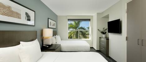 A hotel room with a bed, a desk, a lamp, a TV, a wardrobe, and a window with a view of palm trees.