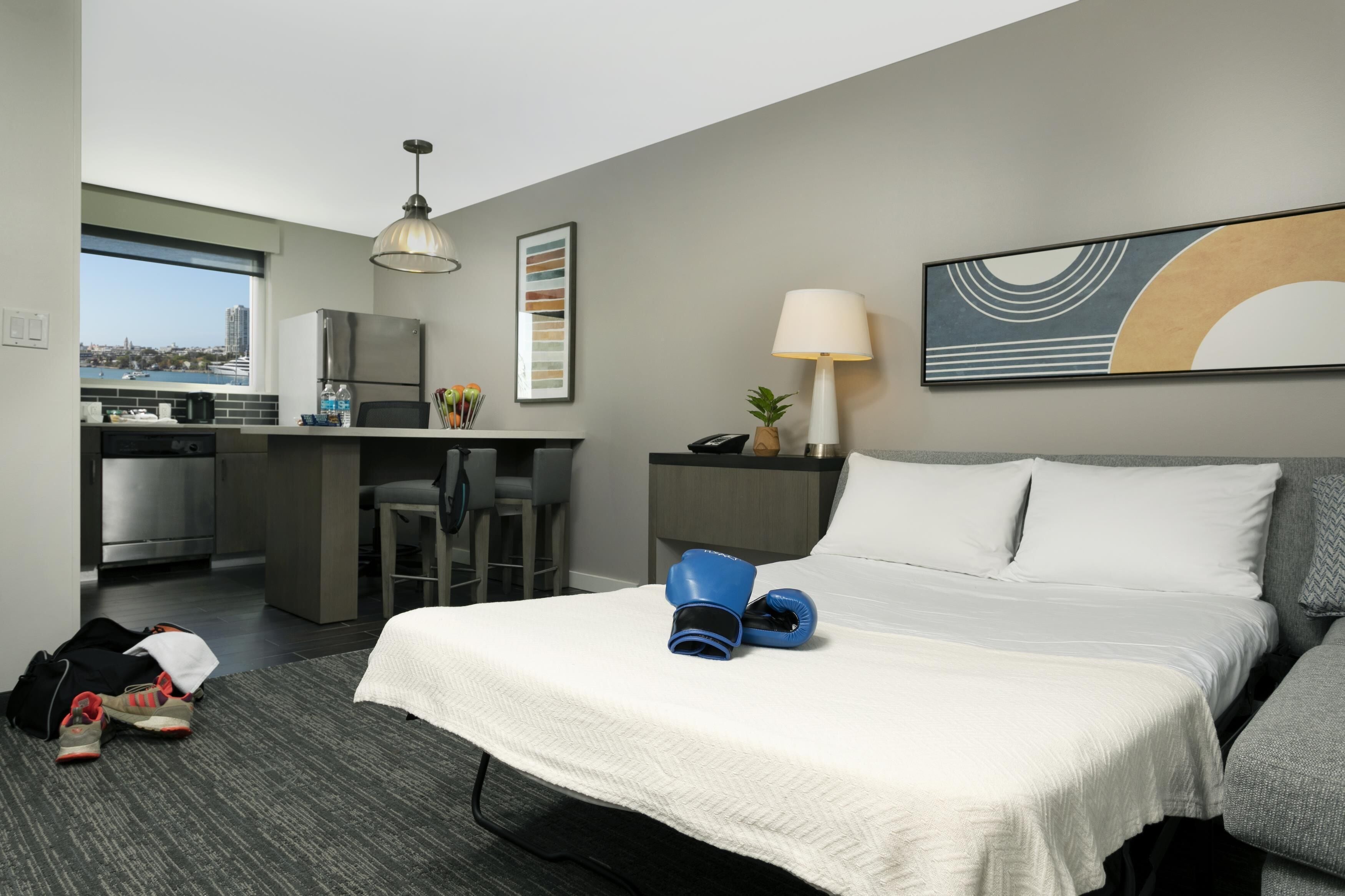 A hotel room with a bed, a desk, a chair, a lamp, and a view of the city.