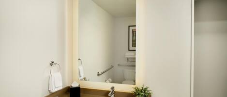 Une salle de bain avec un lavabo blanc, un miroir, un porte-serviettes, une barre d’appui et un tableau encadré au mur.