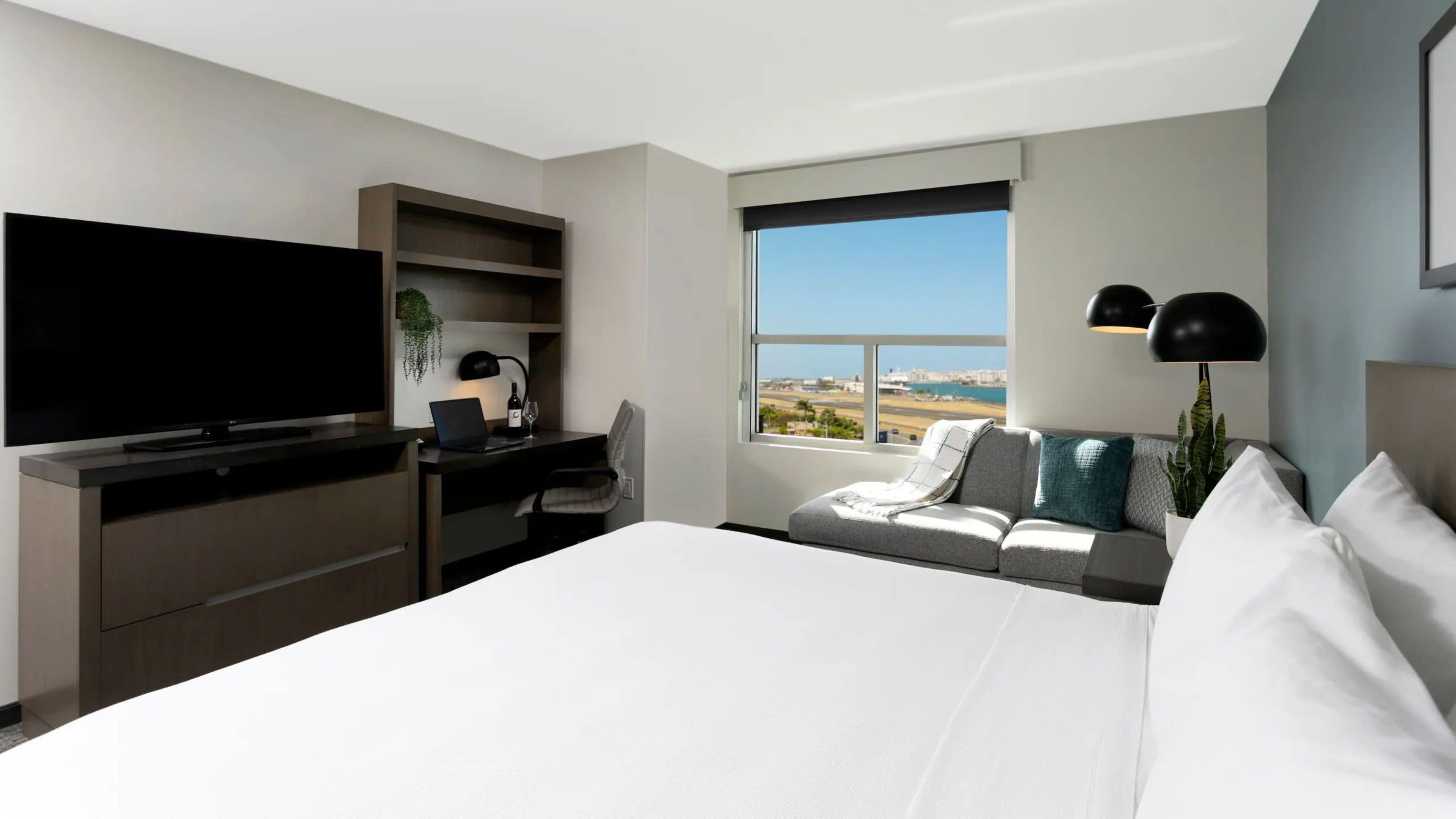Quarto de hotel moderno com uma cama grande, uma escrivaninha com um laptop, uma televisão de tela plana, uma janela com vista para a cidade e um sofá com almofadas.