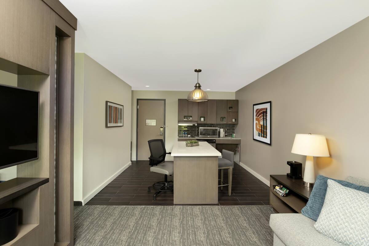 Een moderne hotelkamer met een flatscreen televisie, een bureau met stoel, een kitchenette, een bank met lamp en een lijst aan de muur.