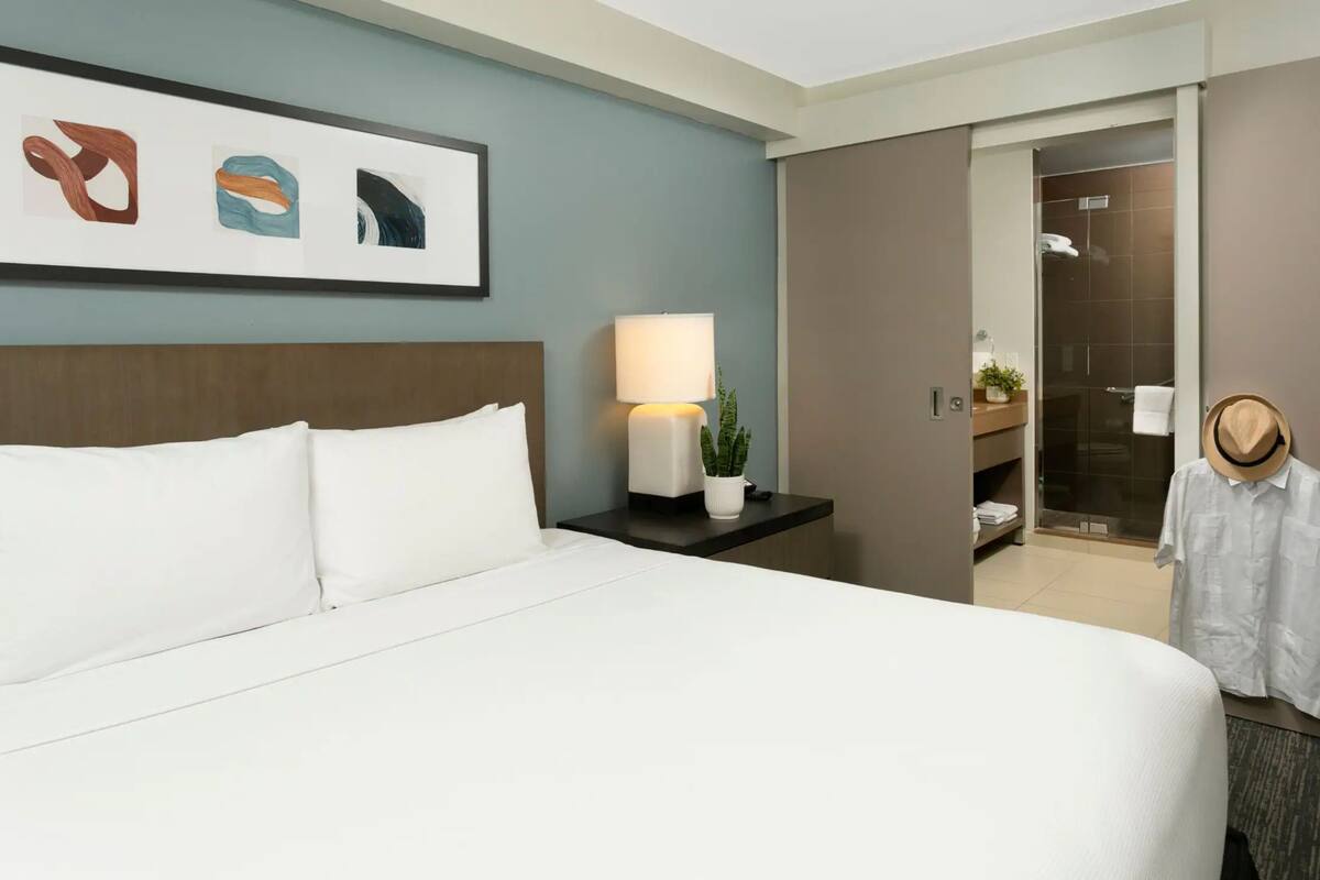 Hotelkamer met een bed, nachtkastje met lamp, een potplant, een ingelijst kunstwerk en een kast met handdoek en hoed.