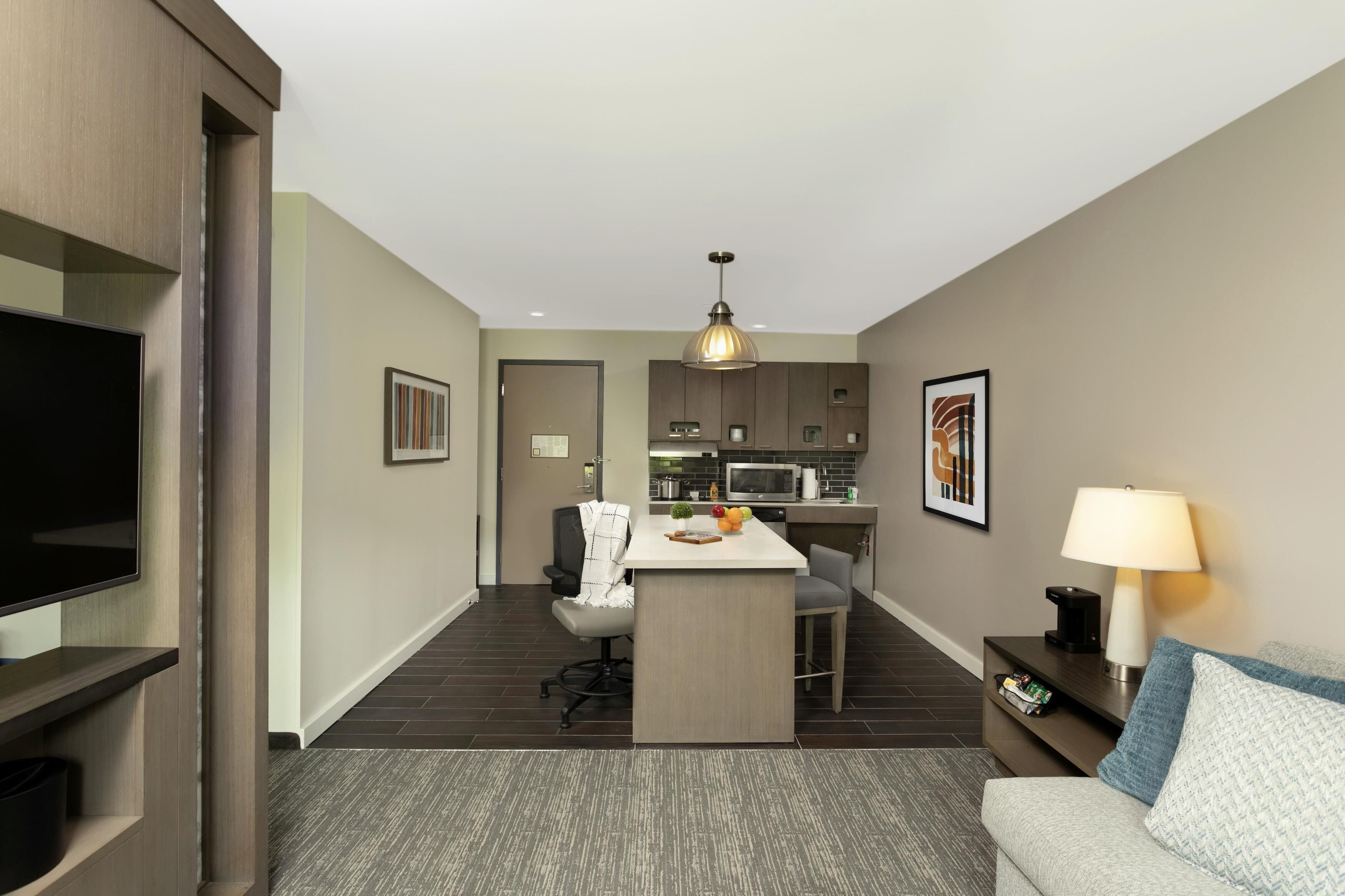 A modern hotel room with a TV, a small kitchen area, a dining table, and a sofa.