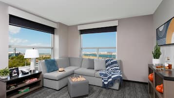 A modern living room with a sectional sofa, a small ottoman, a chess table, a lamp, a bookshelf, and a view of the city.