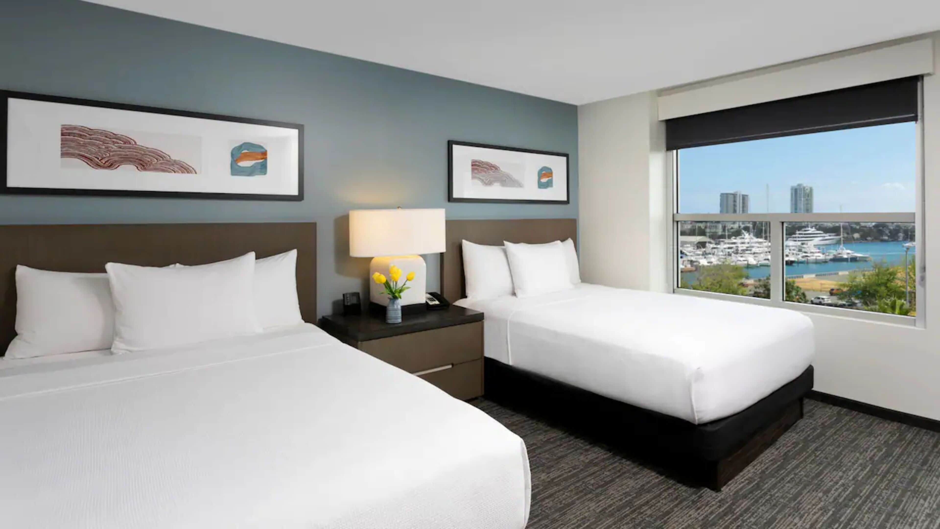 A hotel room with two beds, a nightstand with a lamp, and a window with a marina view.
