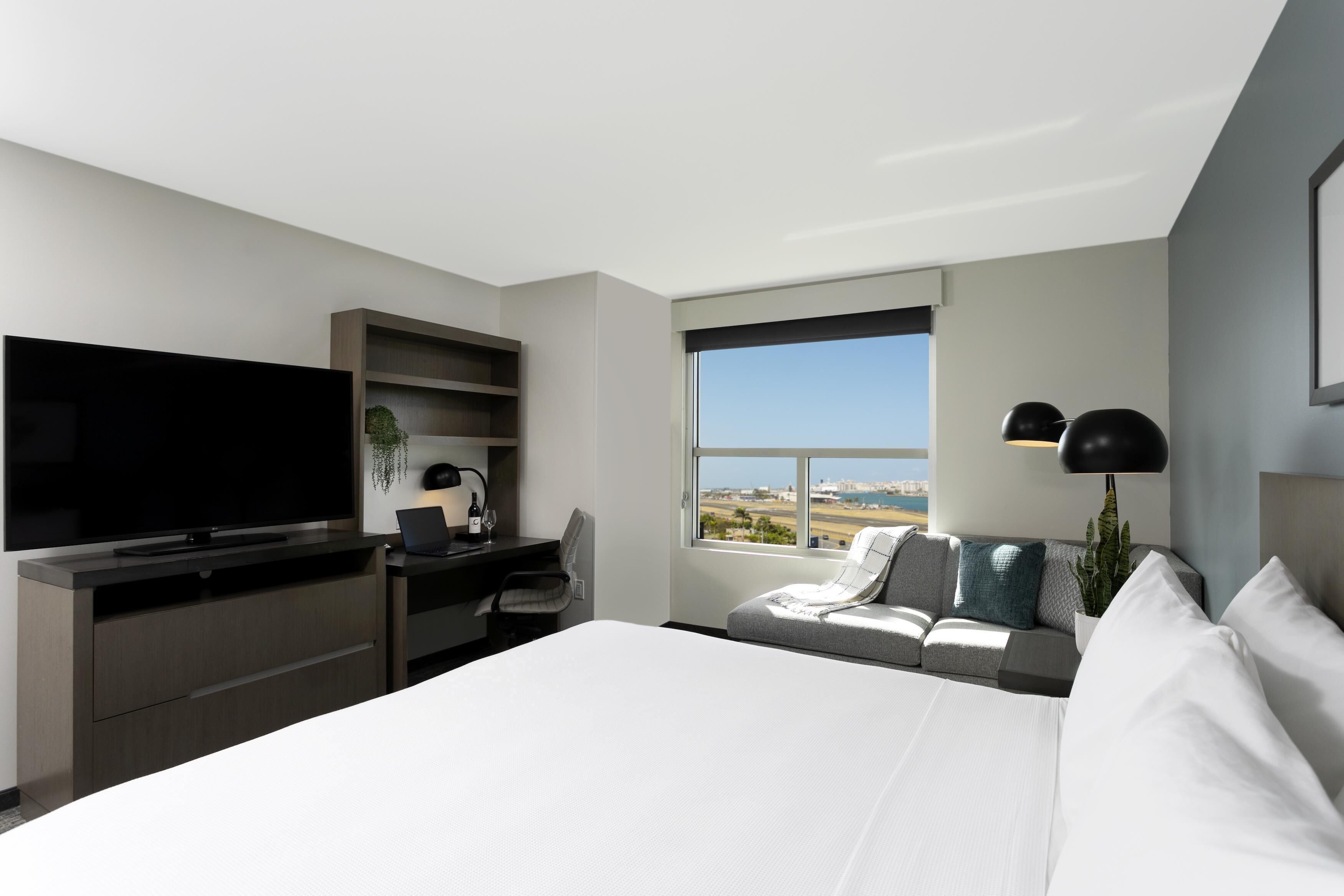 Une chambre d’hôtel moderne dotée d’un grand lit, d’une télévision à écran plat, d’un bureau avec une chaise, d’un canapé et d’une grande fenêtre donnant sur le paysage.