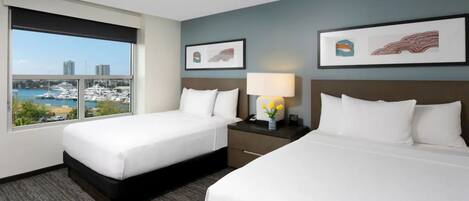 A hotel room with two beds, a window with a marina view, a nightstand with a lamp, and framed artwork on the wall.