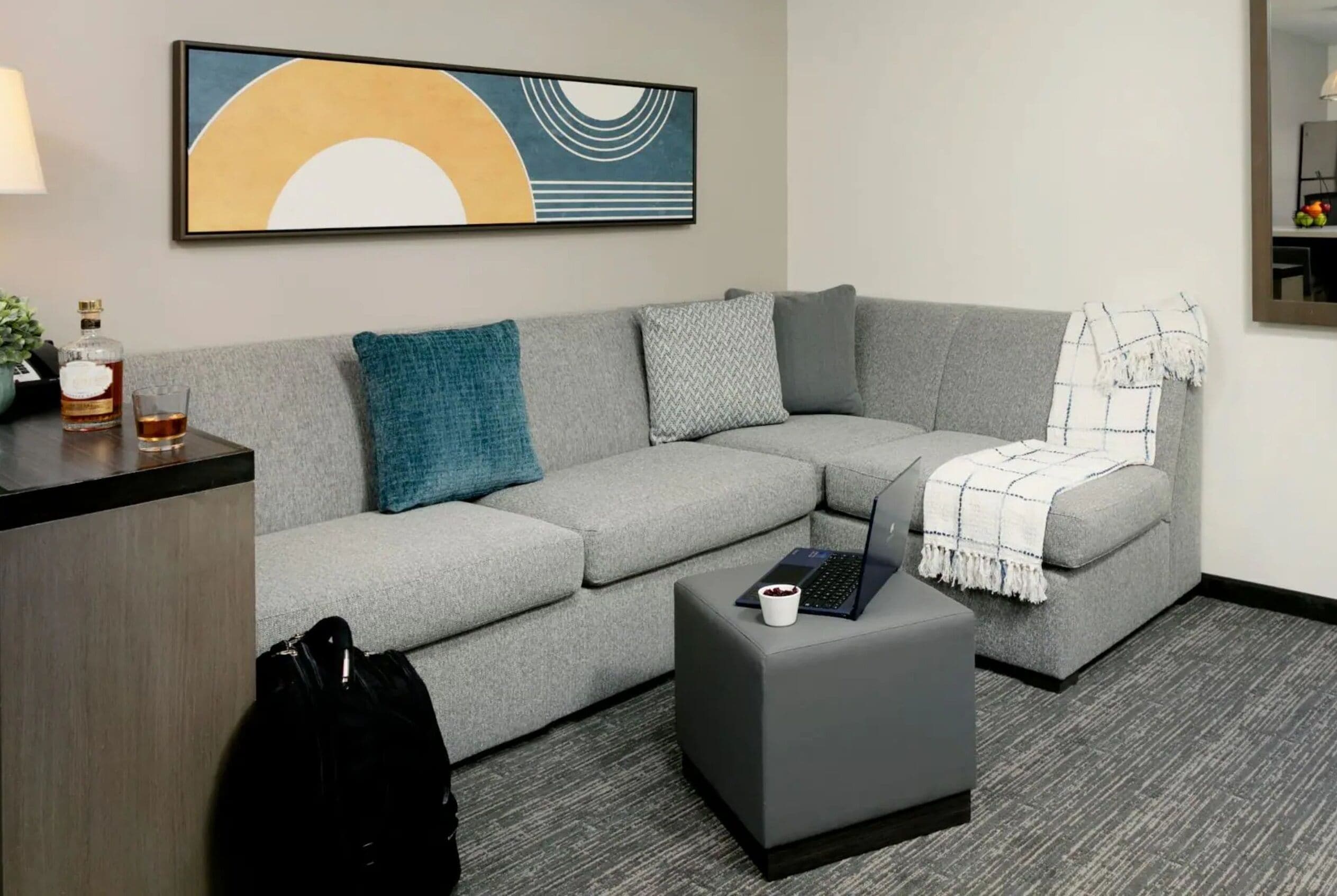 A modern living room with a grey sectional sofa, a small grey ottoman, a laptop, a glass of whiskey, and a decorative abstract painting on the wall.
