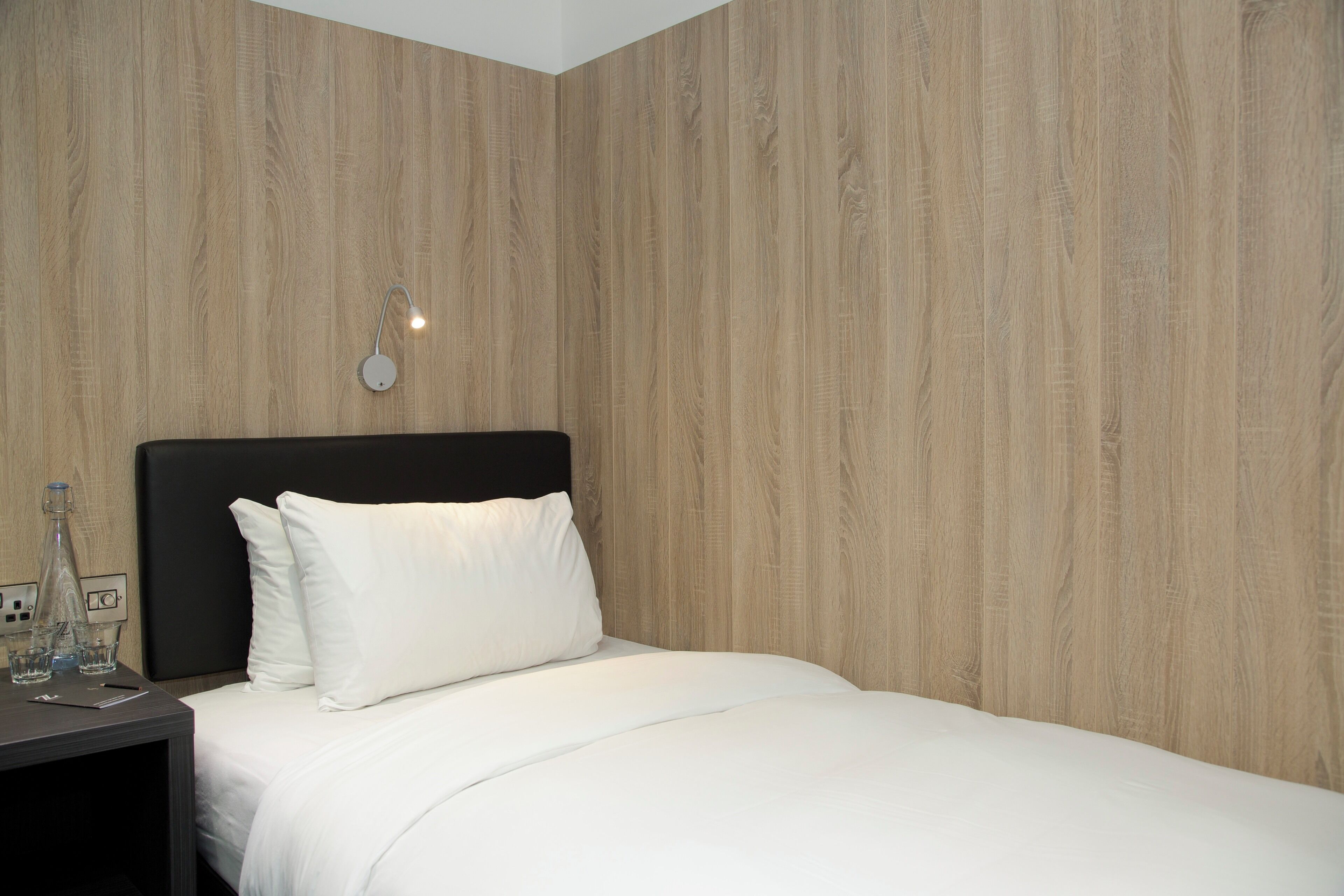 A hotel room with a wooden wall, a black headboard, white bedding, a bedside table with a bottle, glasses, and a phone.