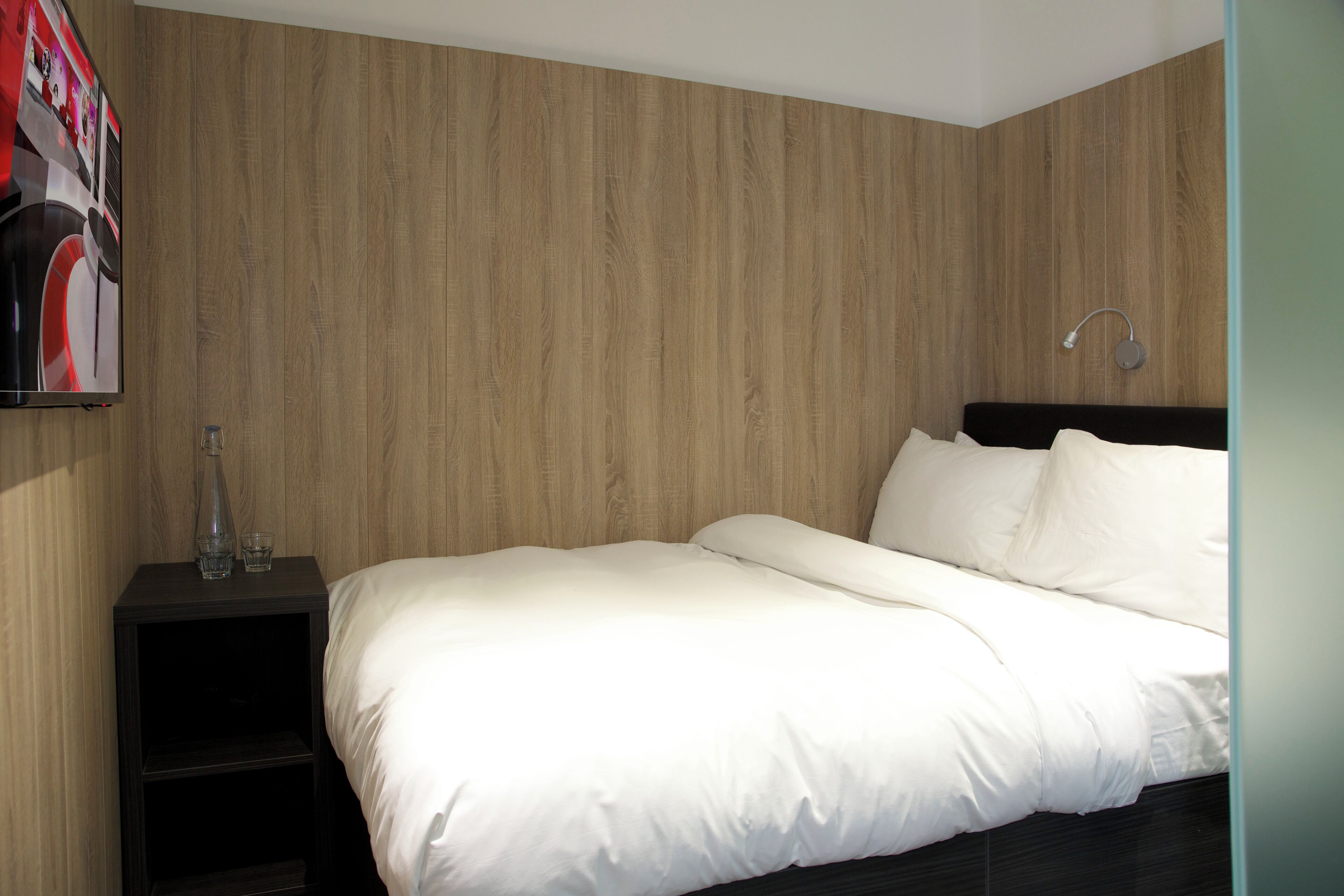 A modern bedroom with a wooden wall, a bed with white bedding, a nightstand with a bottle and glasses, and a wall-mounted light fixture.