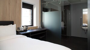 Camera d'hotel moderna con un letto, una scrivania con una macchina del caffè, un box doccia in vetro e vista sulla città.
