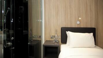 A hotel room with a glass-enclosed shower, a bedside table with a bottle and glasses, and a bed with a white pillow.