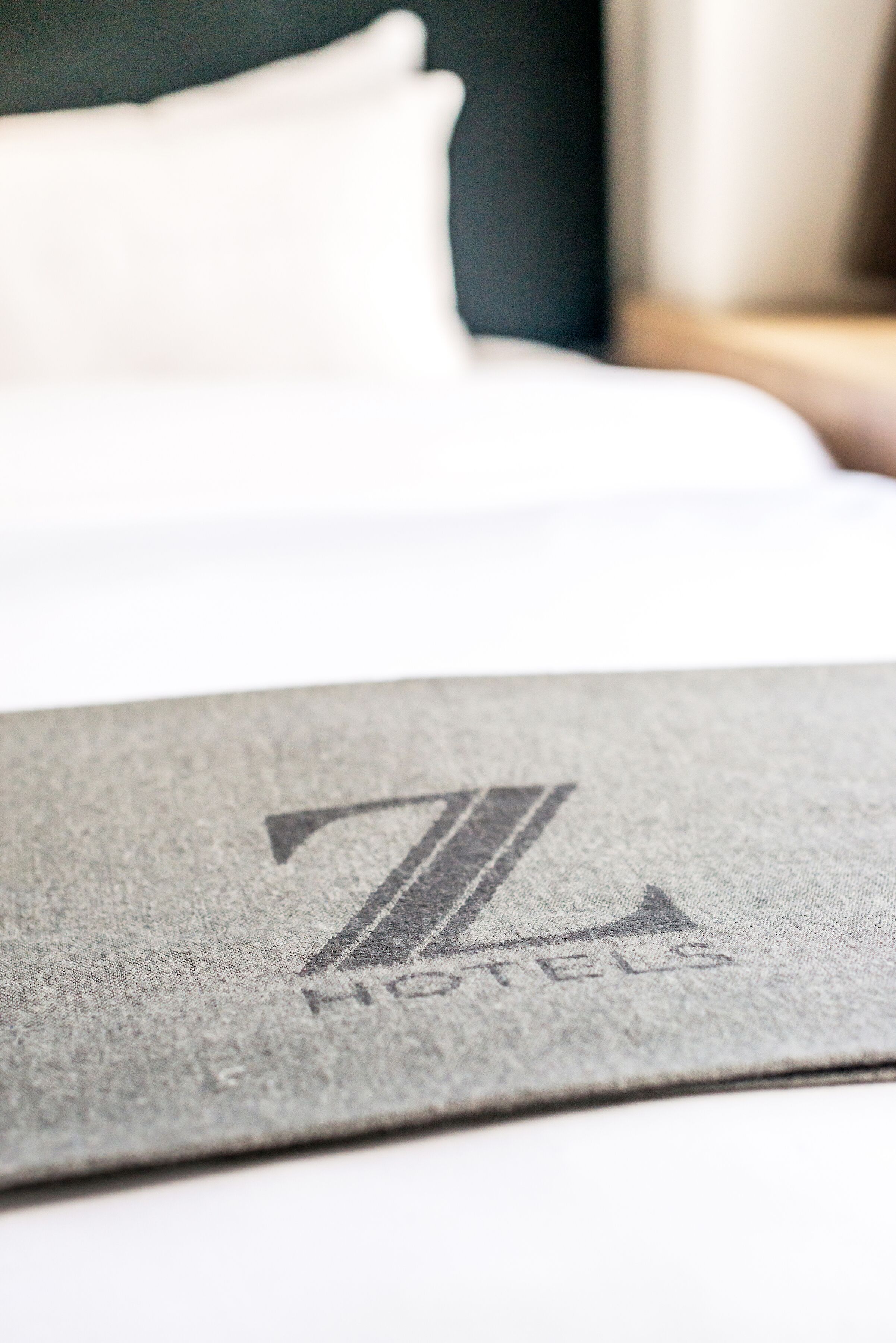 A close-up of a bed with a white pillow and a grey textured cover featuring a logo and the word "HOTELS".
