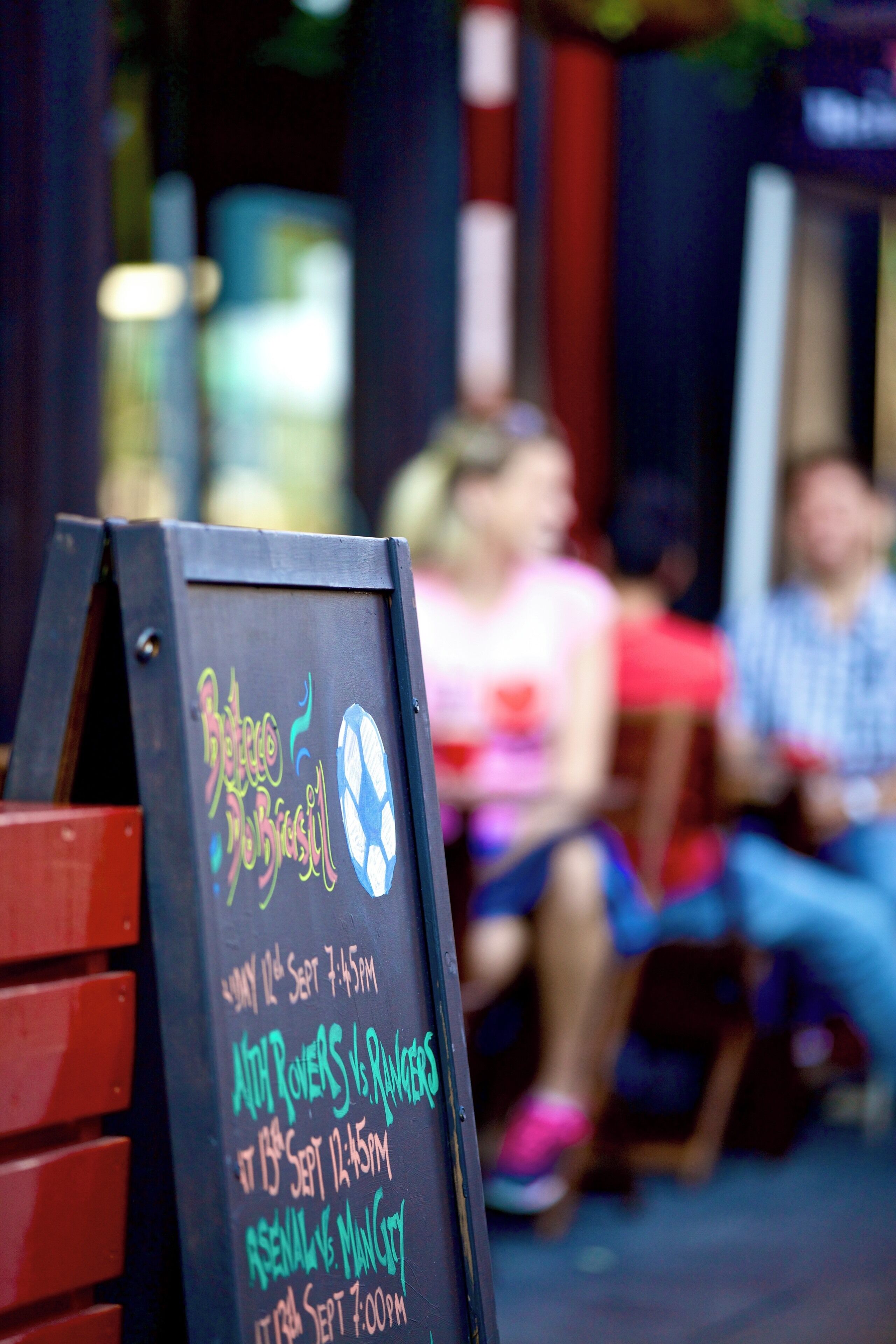 A chalkboard with event details, a red bench, and a blurred background of people.