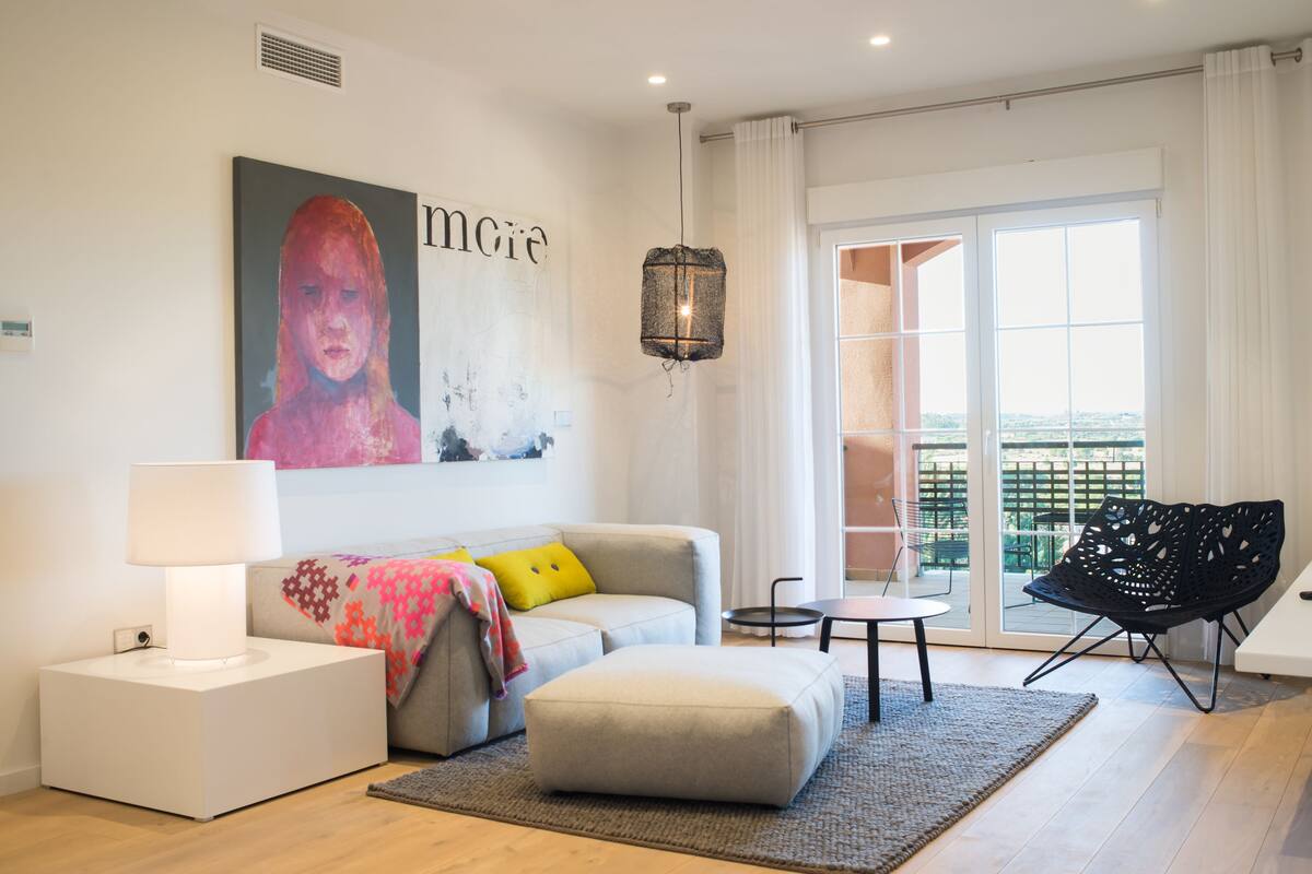 Una sala de estar moderna con un sofá, un reposapiés y una butaca negra. Se observa un ventanal con cortinas, una lámpara colgante y un cuadro en la pared.