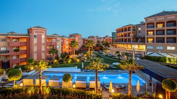 Un hotel amb diversos edificis, una piscina i palmeres.
