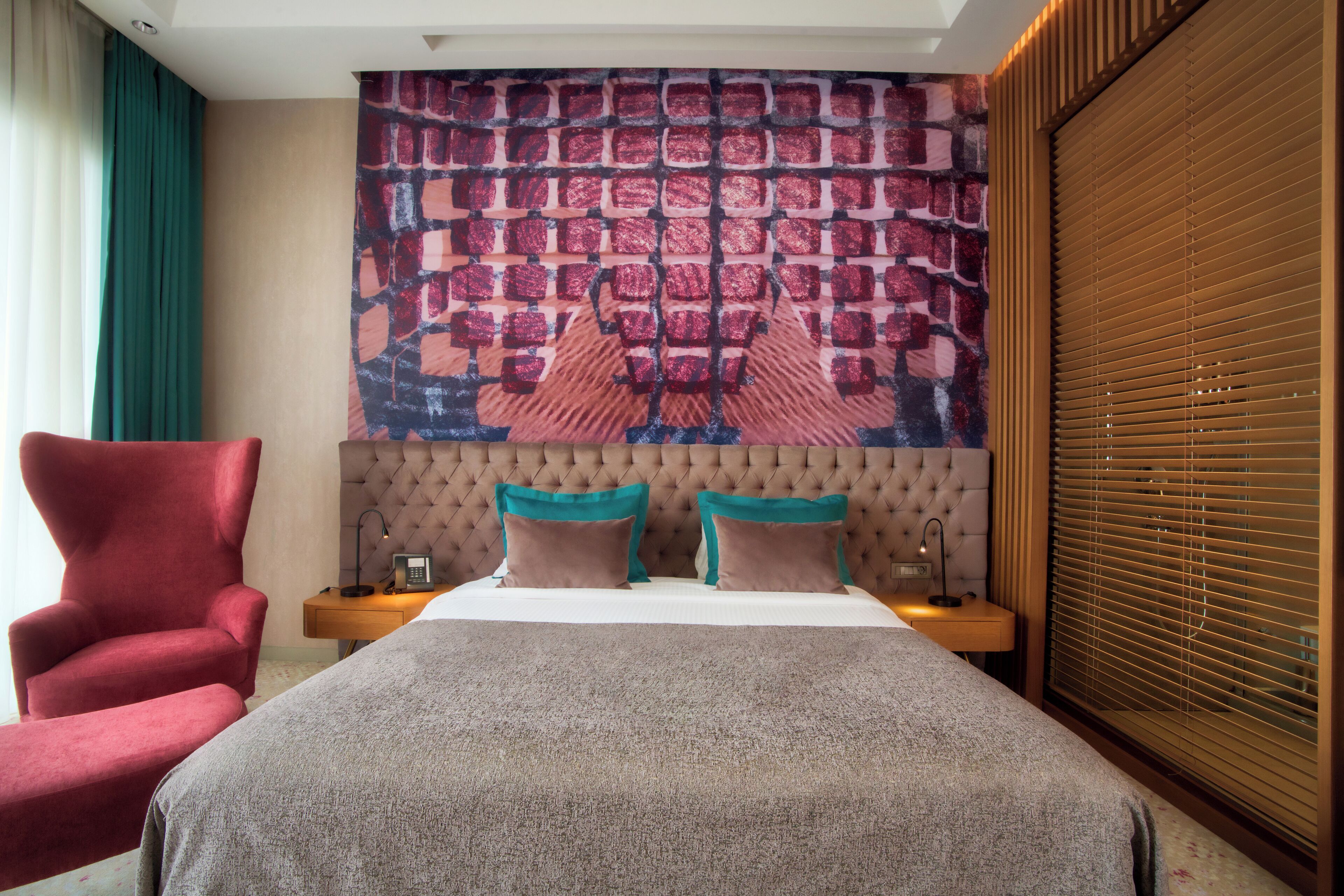 A hotel room with a tufted headboard, a large patterned tapestry, a red armchair, a bedside table with a lamp, and a window with blinds.