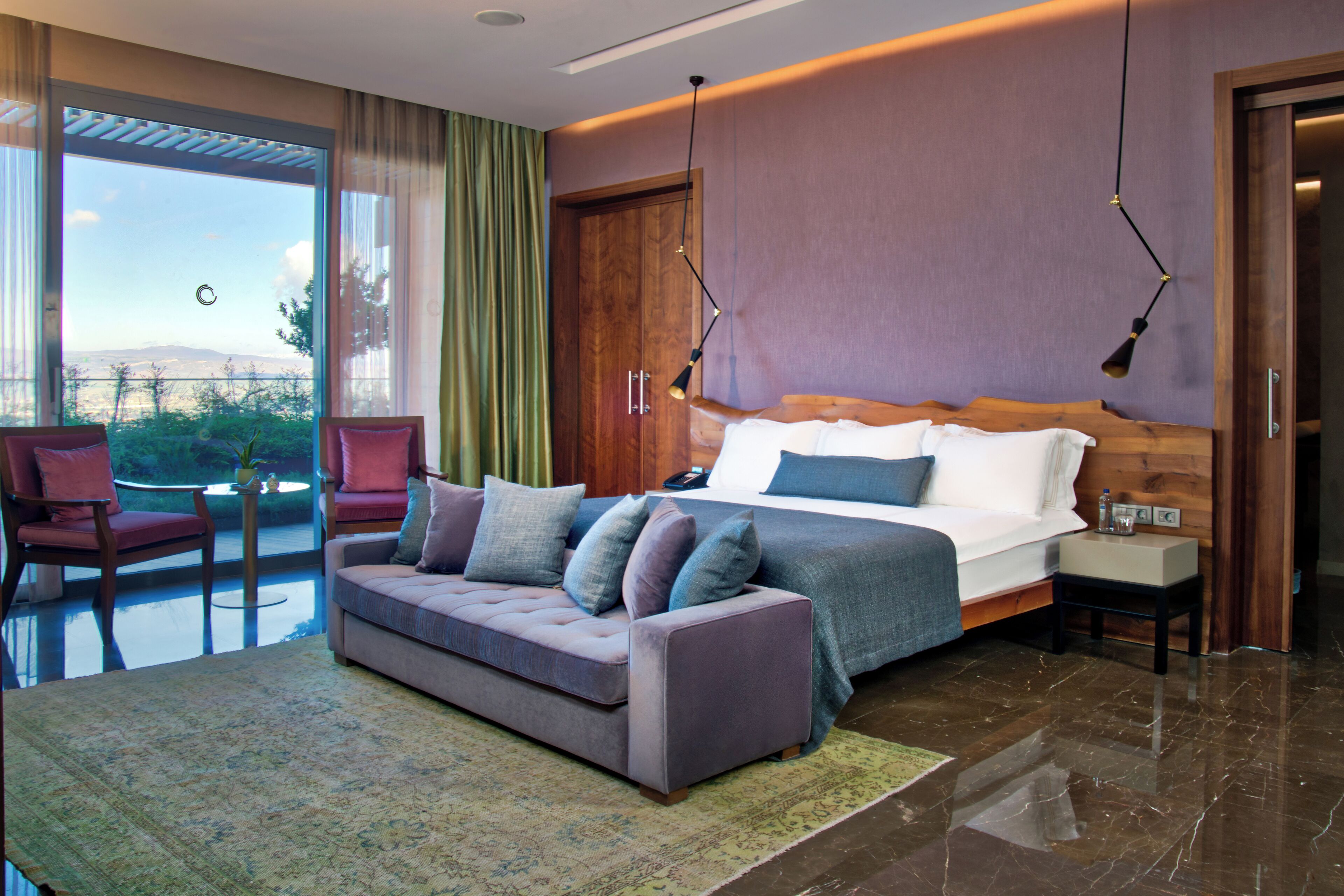 A modern hotel room with a large bed, a sofa, a small table, and a view of the outdoors.