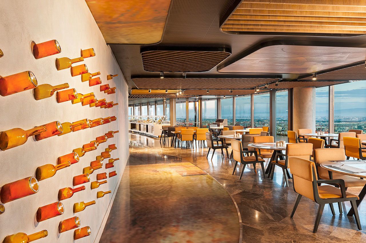 Un restaurante moderno con una larga pared de botellas vacías, amplias ventanas y un suelo pulido.