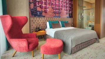 A modern hotel room with a tufted headboard, a large bed, a red armchair, a small ottoman, and a glass-enclosed shower.