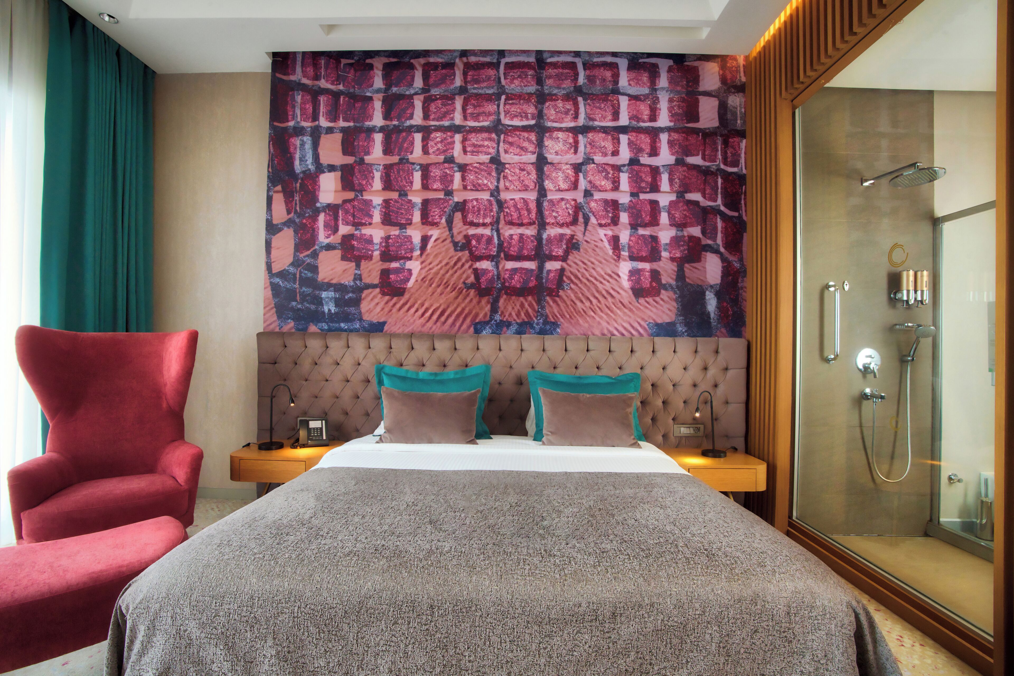 A hotel room with a large bed, a red armchair, a nightstand, a wall tapestry, and a glass-enclosed shower area.