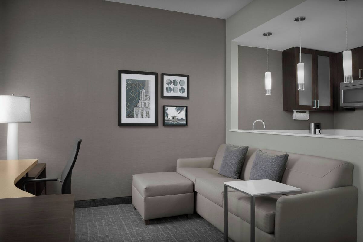 A modern hotel room with a sofa, a small table, a desk, and a kitchenette area.