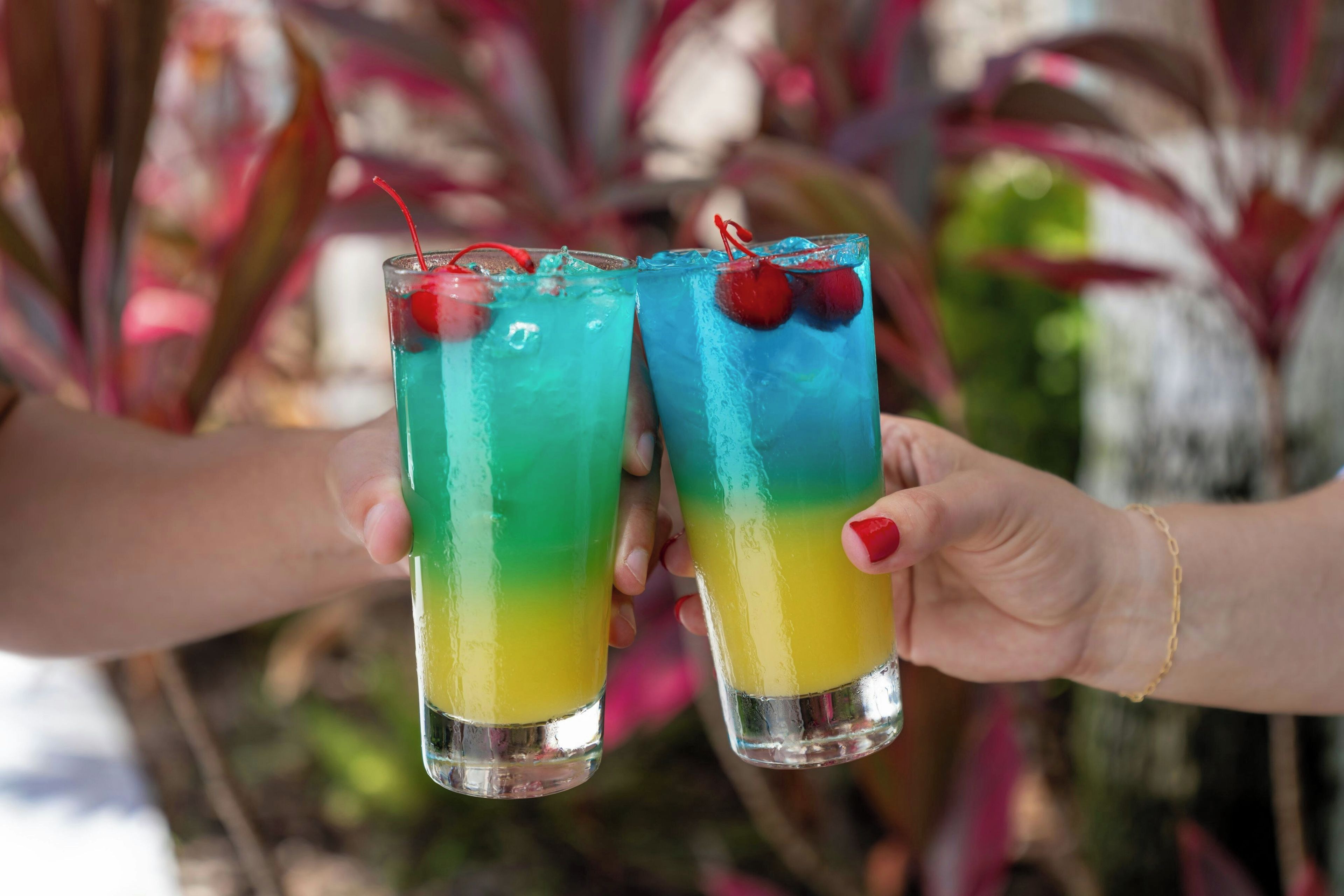 Two glasses of a layered drink with a cherry garnish, held by hands.
