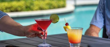 A poolside setting with a table holding two drinks, one in a martini glass and the other in a short glass, with a pair of sunglasses and a magazine on the table.