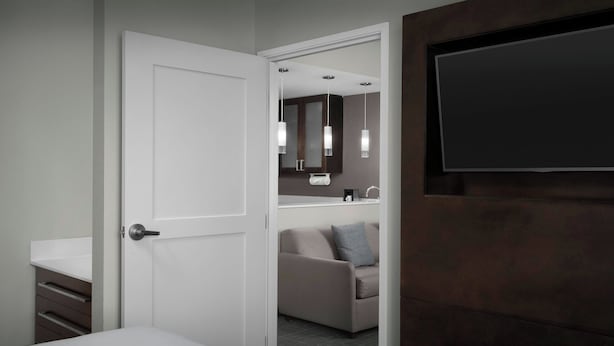 Habitación de hotel con cama, baño con espejo, lavamanos y televisor en la pared.