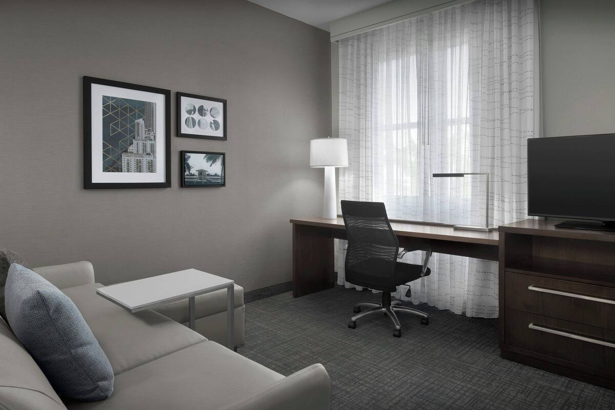 A hotel room with a sofa, a desk with a computer, a TV, and framed pictures on the wall.