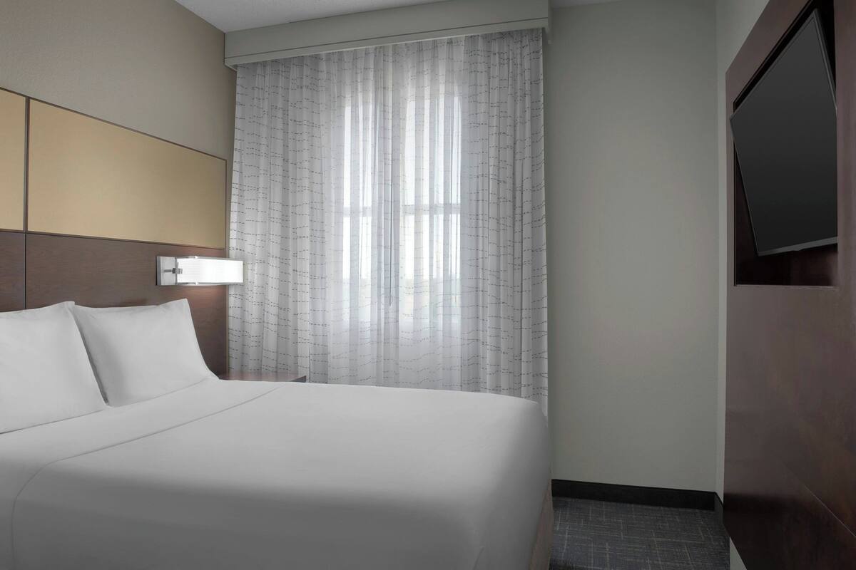 A hotel room with a large bed, a headboard, a window with sheer curtains, and a flat-screen TV mounted on the wall.