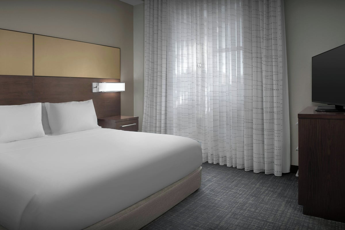 A hotel room with a large bed, a nightstand, a TV, and a window with sheer curtains.