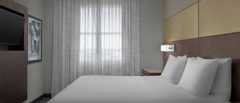 Habitación de hotel con una cama grande, un televisor montado en la pared, una mesita de noche y una ventana con cortinas translúcidas.