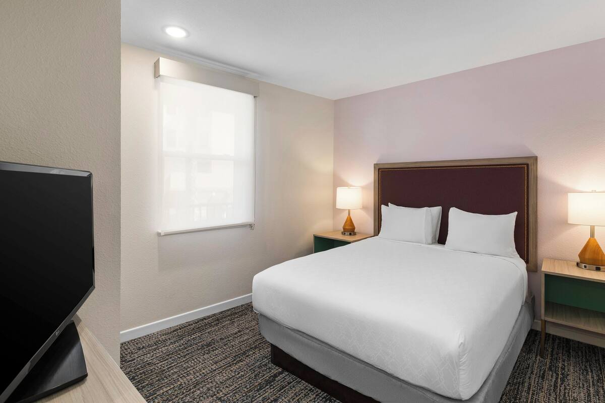 A hotel room with a large bed, two bedside tables with lamps, a flat-screen TV, and a window with blinds.