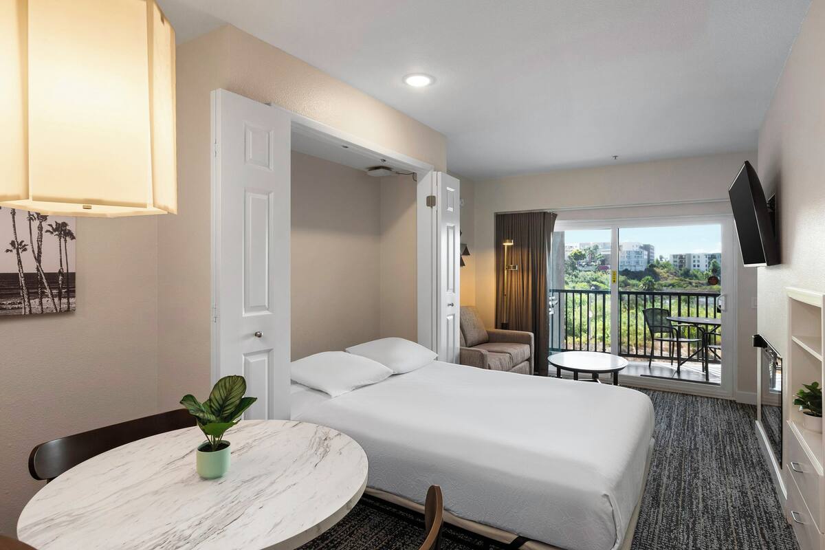 A hotel room with a bed, a dining table with a plant, a balcony with a view, and a TV mounted on the wall.