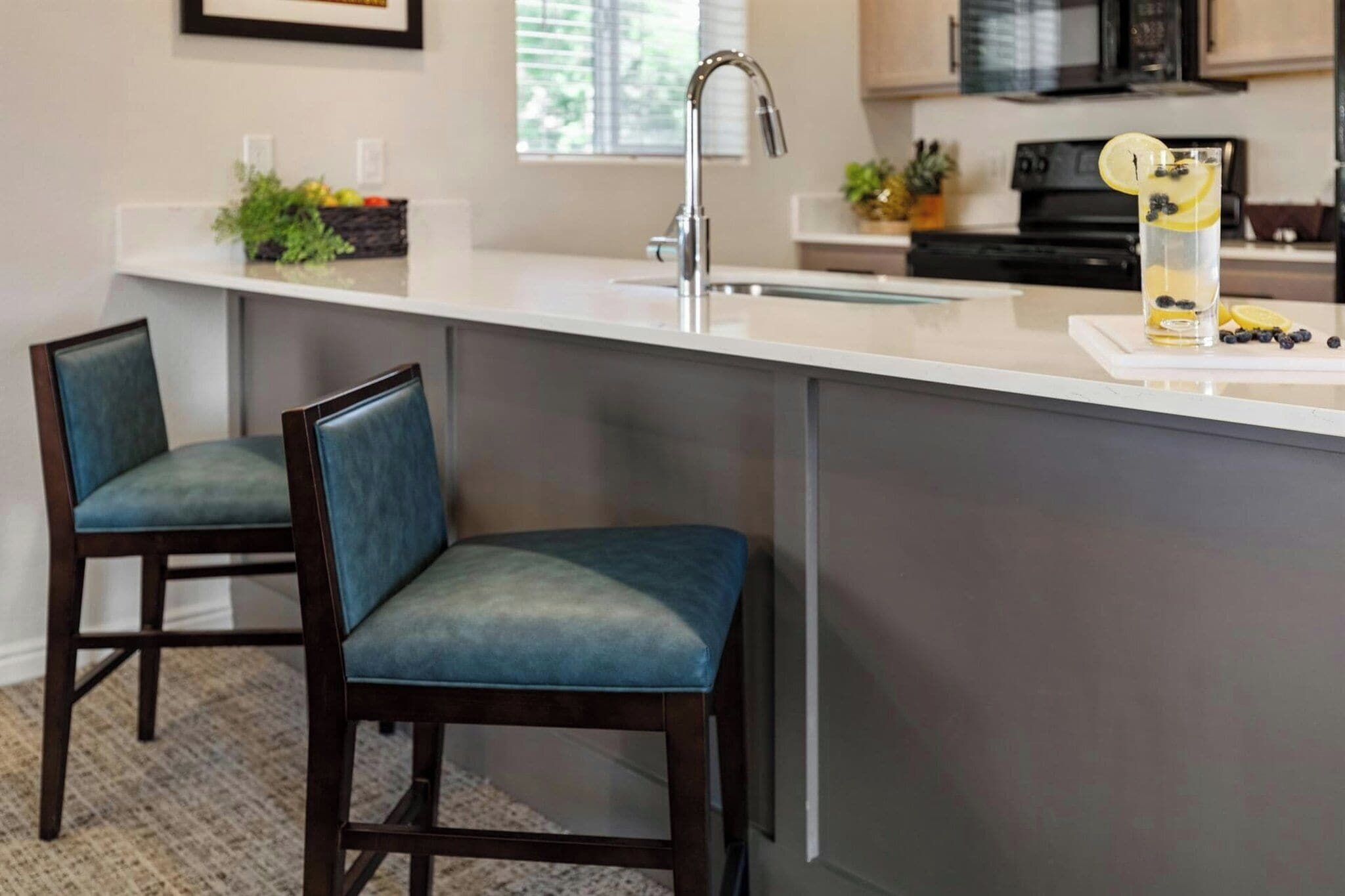 A kitchen with a central island, two bar stools, a stainless steel faucet, and a glass of beverage with lemon and blueberries.