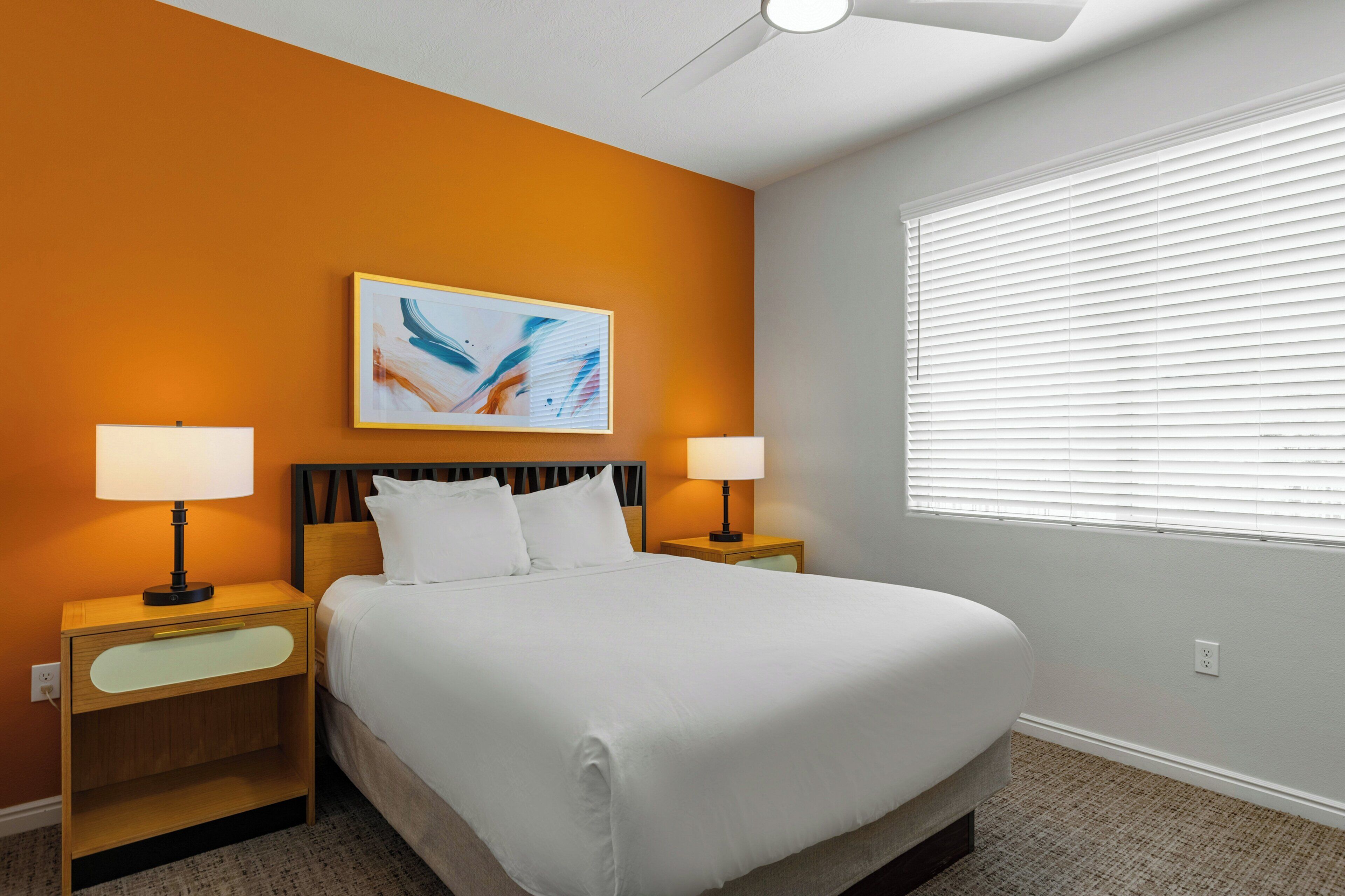 A hotel room with a bed, two bedside tables with lamps, a ceiling fan, a window with blinds, and a painting on the wall.